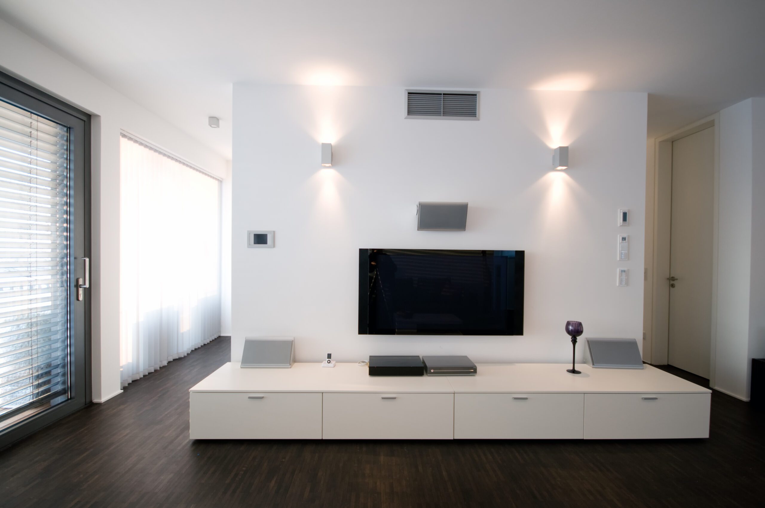 A modern living room features a wall-mounted flat-screen TV with a minimalist white media console below, illuminated by two wall-mounted lights. Vertical blinds cover a large window beside them.